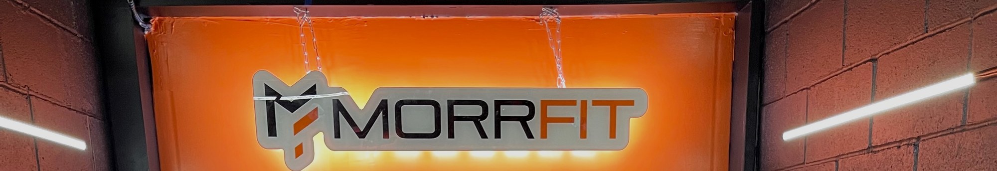 The lit MORRFIT sign on the orange entry wall, with LED strips on the brick